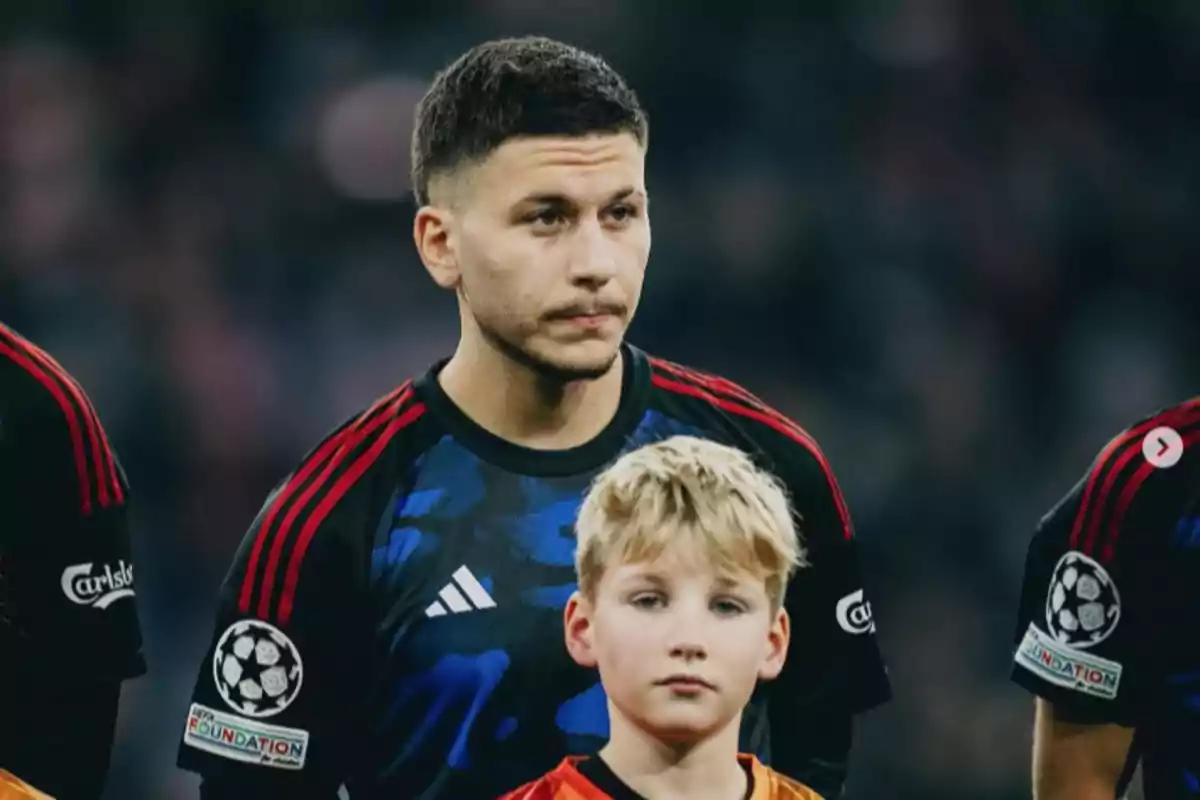 A player in a black and red uniform is standing on the field next to a blond boy before a match.