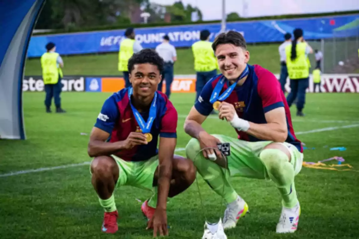 Dos futbolistas jóvenes con uniforme del Barcelona posan sonrientes en el campo mostrando sus medallas de oro tras un partido
