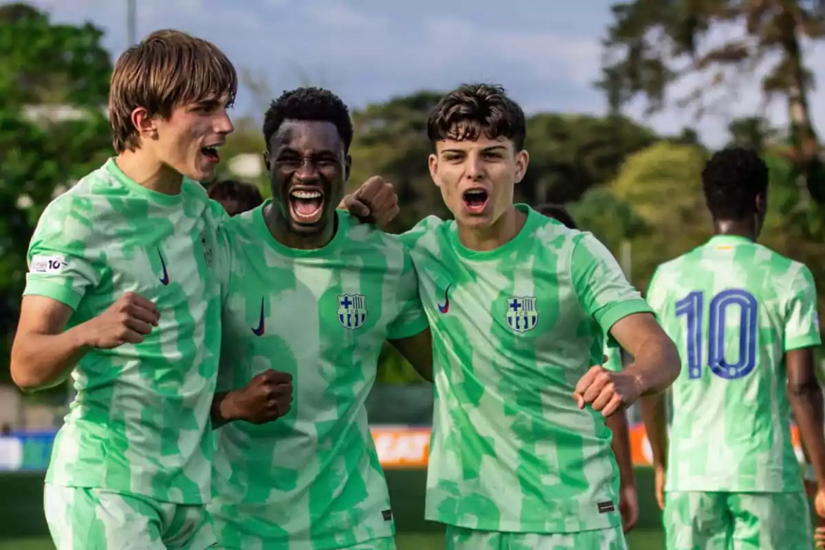 Three young players celebrate together on the field wearing green uniforms with blue and white details.
