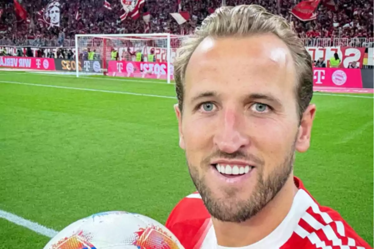 Futbolista sonriente con uniforme rojo sosteniendo un balón en un estadio lleno de aficionados
