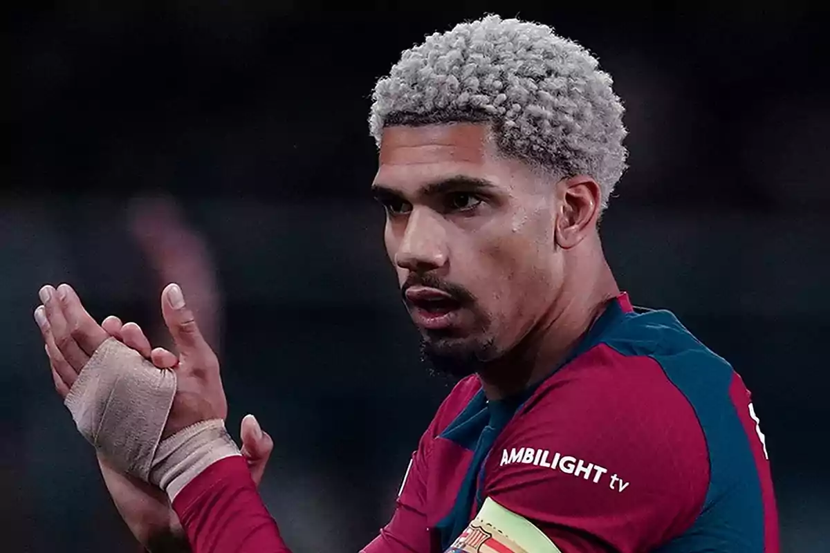 Futbolista con el cabello rizado y teñido de rubio, usando un uniforme azul y rojo, aplaudiendo con una mano vendada. Futbolista con el cabello rizado y teñido de rubio, usando un uniforme azul y rojo, aplaudiendo con una mano vendada.