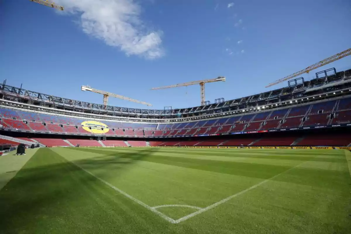 Estadio de fútbol con gradas vacías, césped bien cuidado y varias grúas de construcción alrededor bajo un cielo despejado