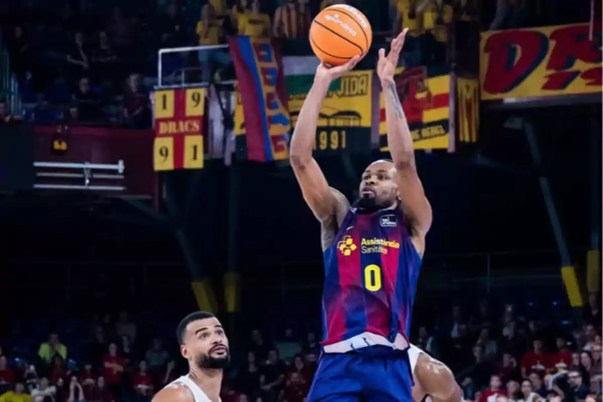 Jugador de baloncesto del Barcelona lanzando el balón durante un partido con público y banderas en el fondo Jugador de baloncesto del Barcelona lanzando el balón durante un partido con público y banderas en el fondo