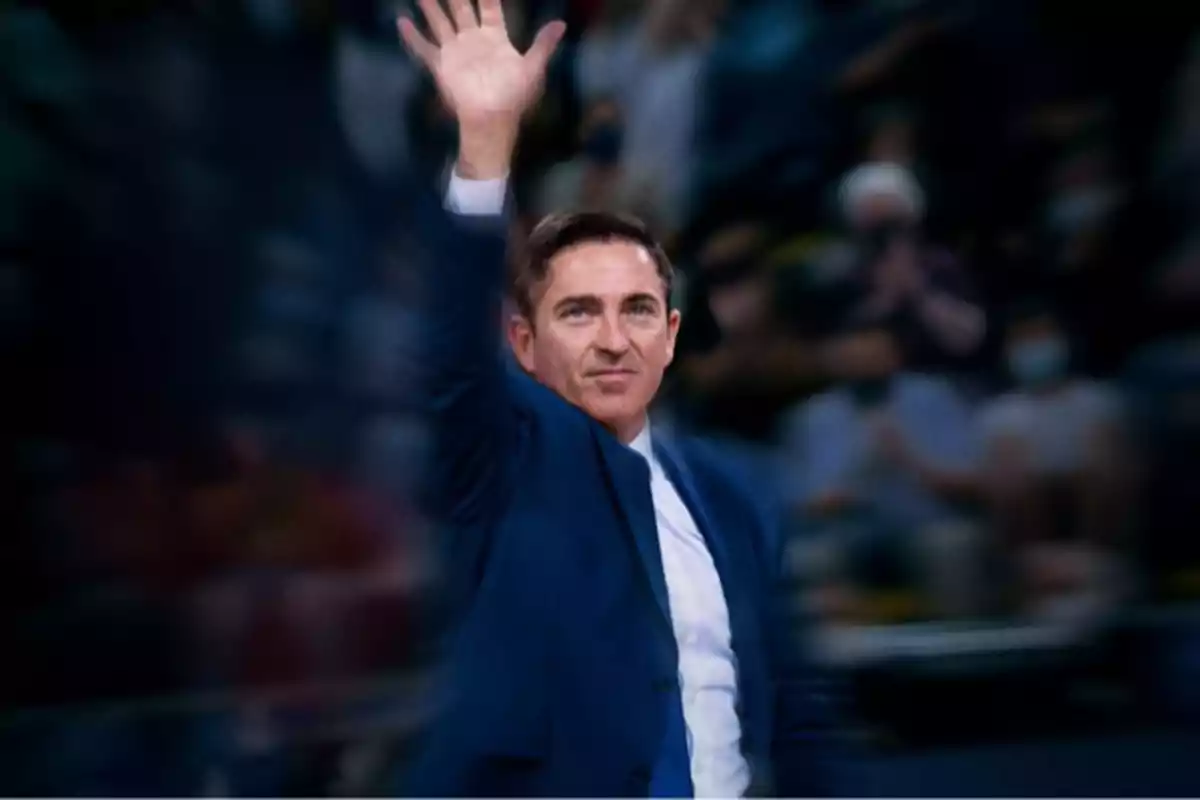 Hombre con traje azul saludando con la mano levantada en un evento deportivo con público desenfocado al fondo Hombre con traje azul saludando con la mano levantada en un evento deportivo con público desenfocado al fondo