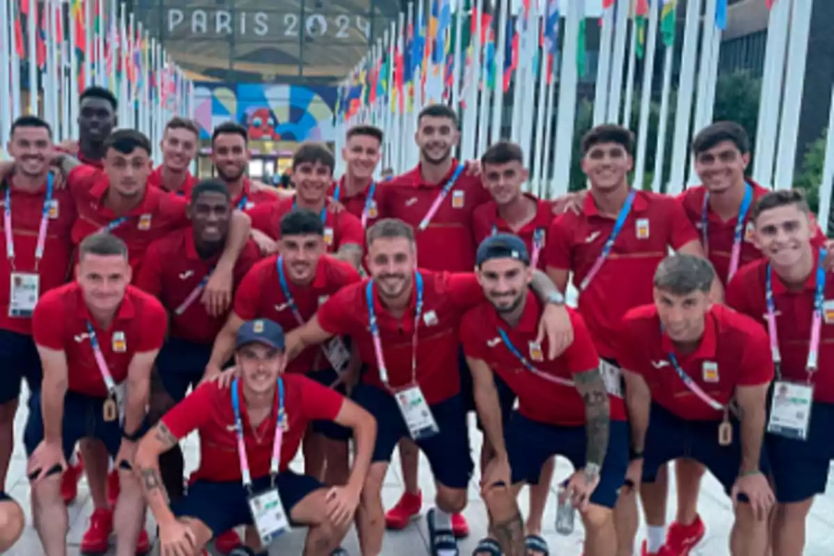 Un grupo de jóvenes deportistas con uniformes rojos posando frente a una entrada decorada con banderas y un cartel que dice 