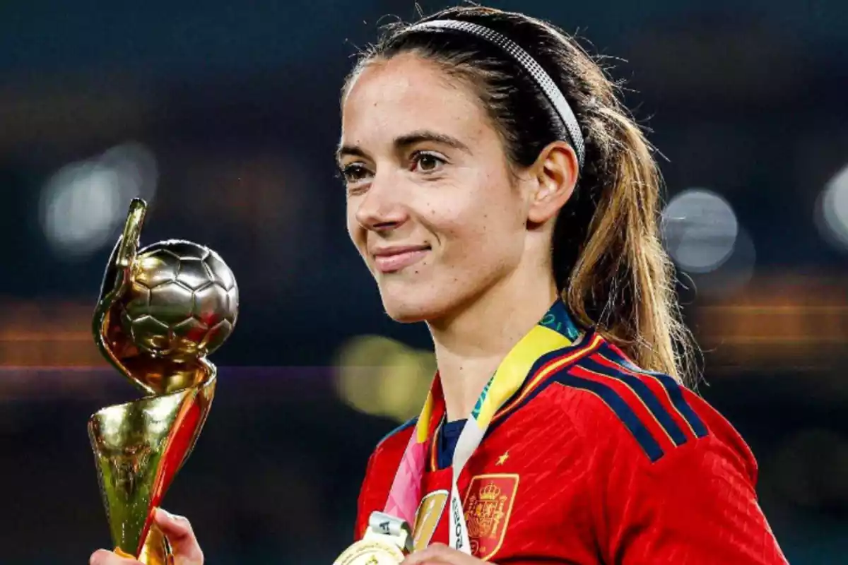 aitana sonriendo mientras lleva una camiseta roja de la seleccion española mientras sujeta un trofeo