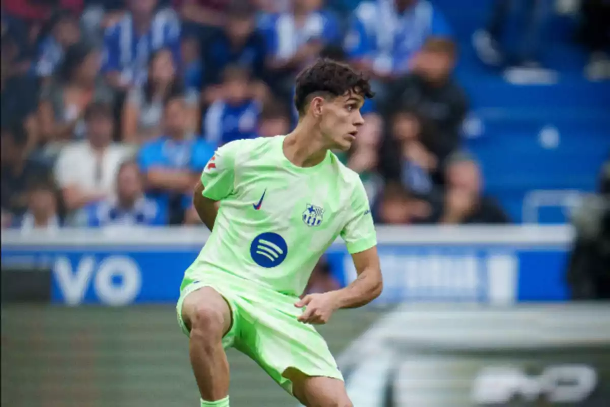 Jugador de fútbol en acción durante un partido, vistiendo un uniforme verde claro con logotipos visibles, en un estadio con público de fondo.