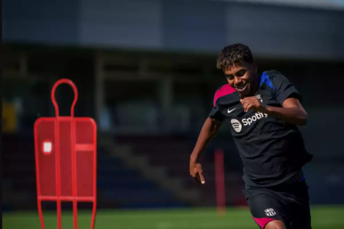 A football player training on the field with a black jersey and a red training dummy in the background.