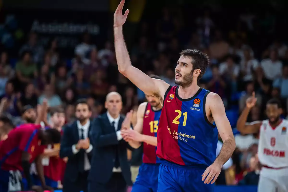 Jugador de baloncesto en uniforme azul y rojo levantando la mano durante un partido con el público y otros jugadores al fondo.