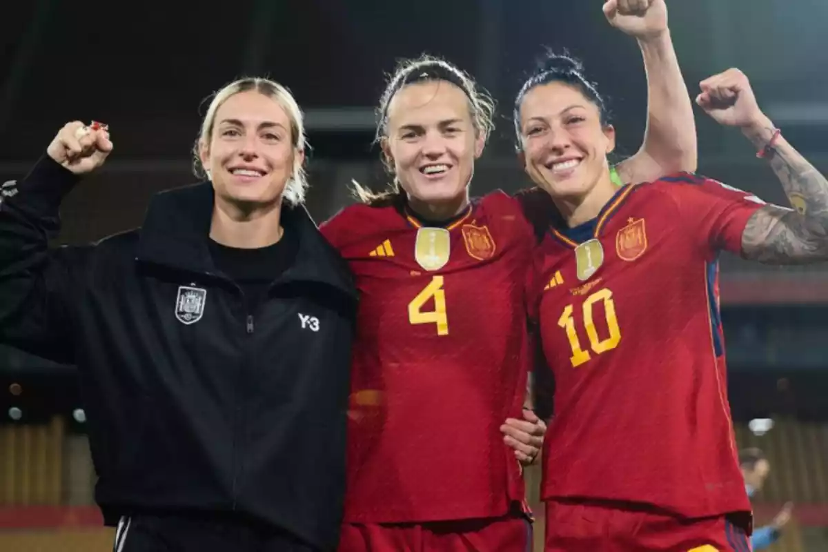Se pueden ver a tres jugadoras, dos vestidas con la equipacion de la selección española y otra vestida de calle. Las tres están abrazadas y con un gesto de alegría.