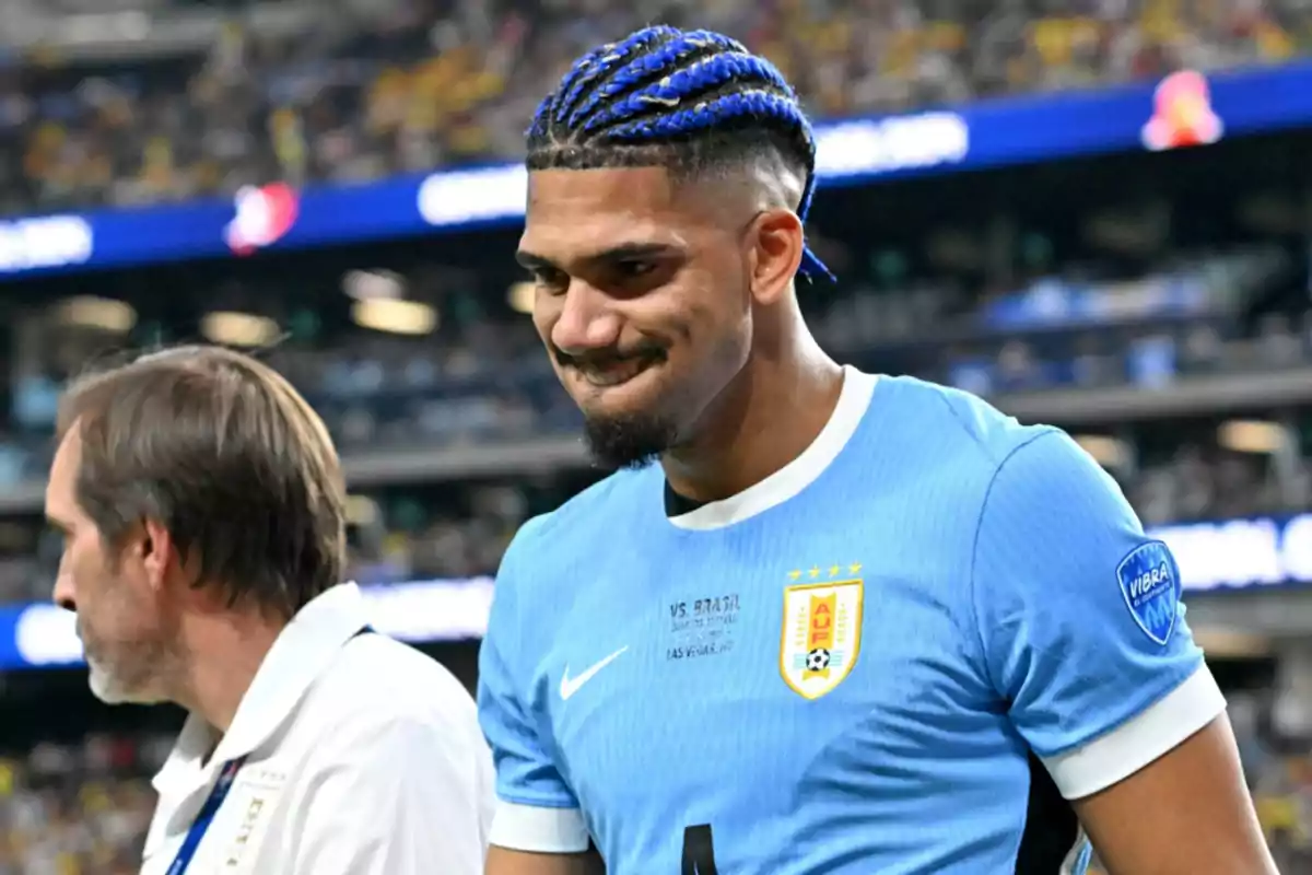 Araujo con trenzas azules y camiseta celeste de la selección de Uruguay, acompañado por otra persona en un estadio lleno de espectadores.