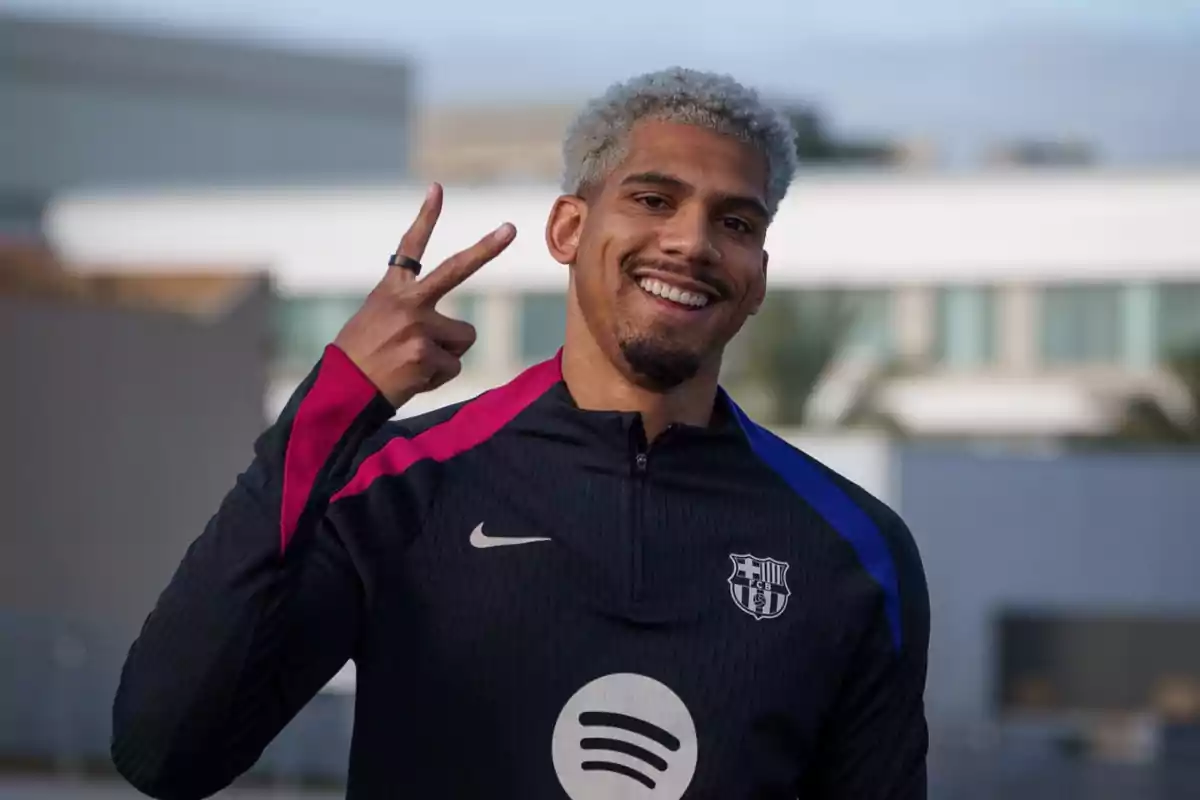 A man with short curly hair smiles as he makes a peace sign with his hand, wearing a black sports jacket with pink and blue details and a logo on the chest.