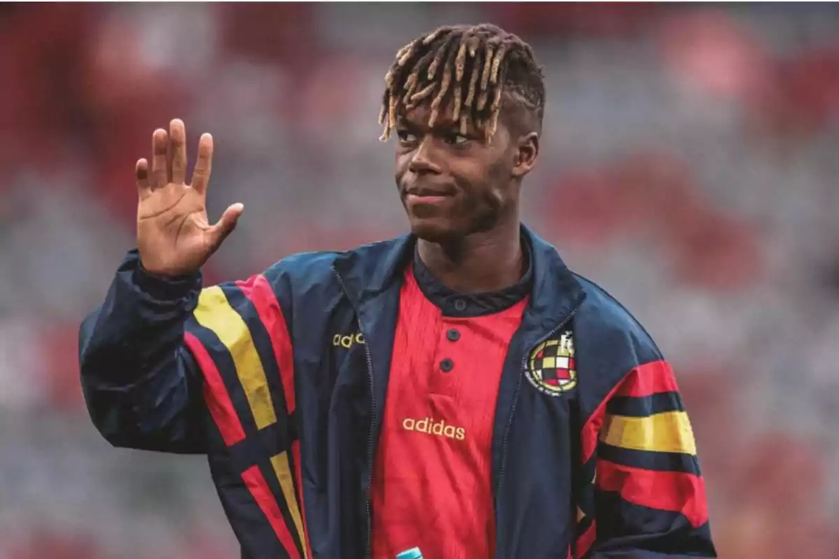 Un jugador de fútbol con una chaqueta azul con detalles en rojo, amarillo y negro, y una camiseta roja, saluda con la mano levantada.