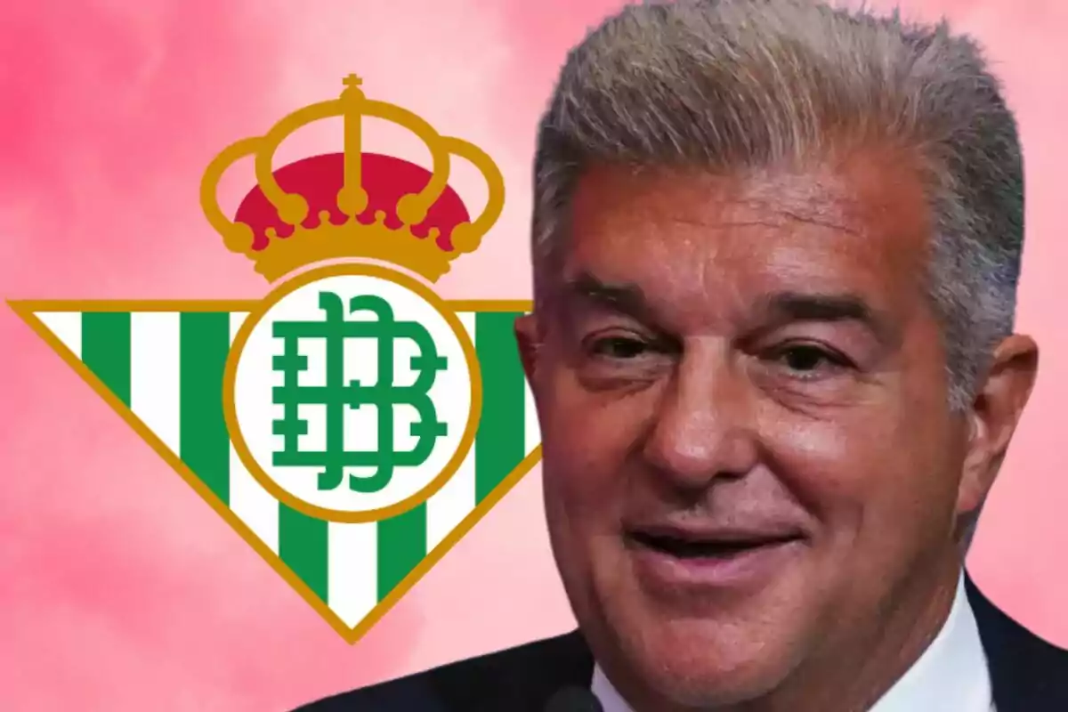 Un hombre sonriente frente al escudo del Real Betis con un fondo rosa.