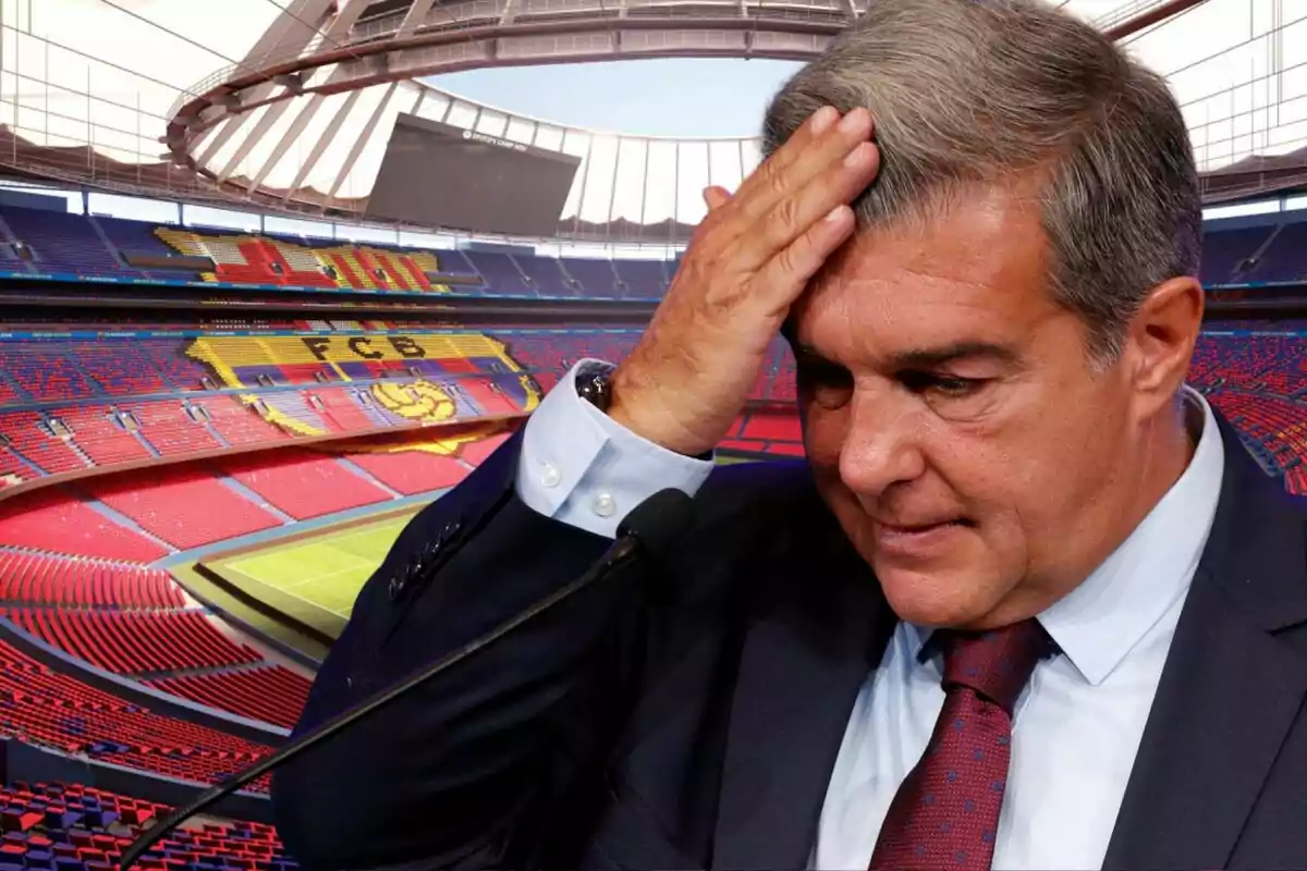 A man in a suit and tie touches his head while standing in front of a microphone with an empty soccer stadium in the background.