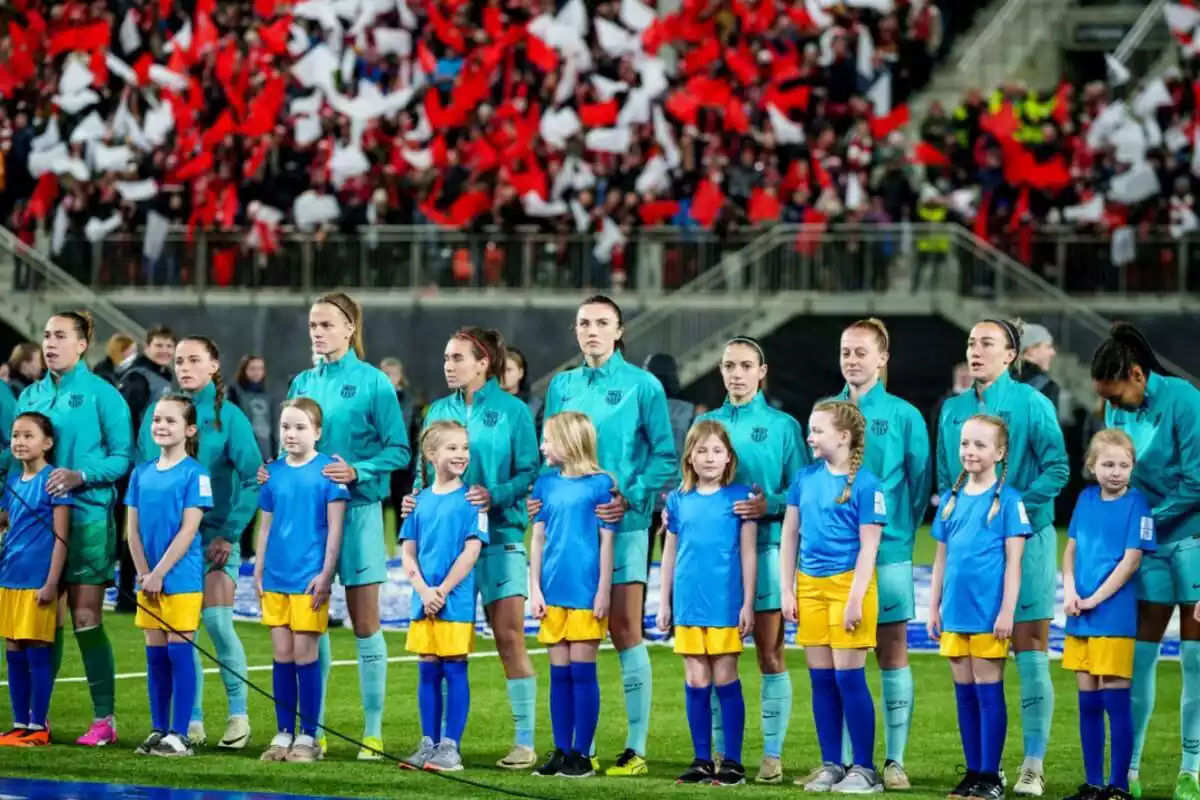 El equipo femenino del FC Barcelona, con indumentaria celeste, preparadas para un partido con el mosaico de la afición rival detrás de ellas
