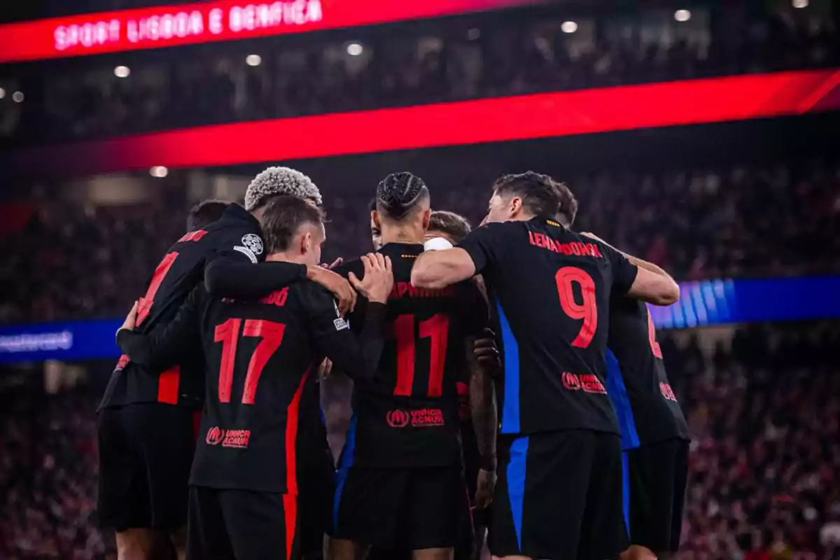 Soccer players in black and red uniforms embrace on the field during a match in a floodlit stadium.