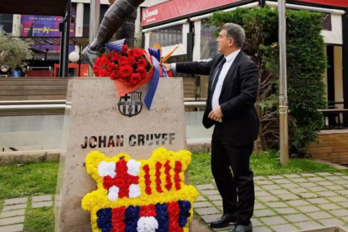laporta frente a una estatua de johan cruyff. el va vestido con un traje negro, corbata negra y camisa blanca. en la pate frontal se ve un escudo de barsa hecho a base de flores.