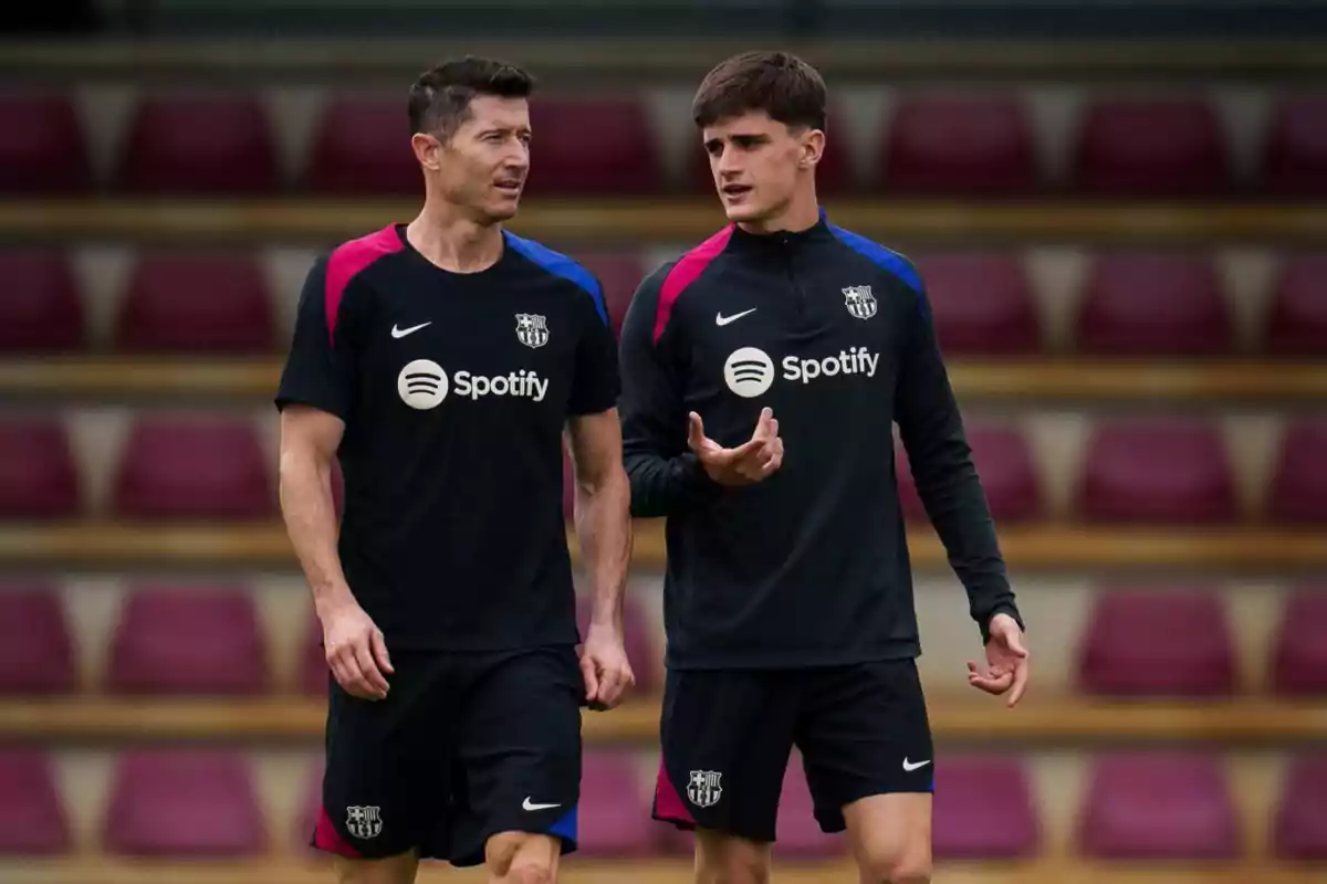 Two FC Barcelona football players, dressed in black training clothes with the Spotify logo, chat as they walk through a stadium with red seats.