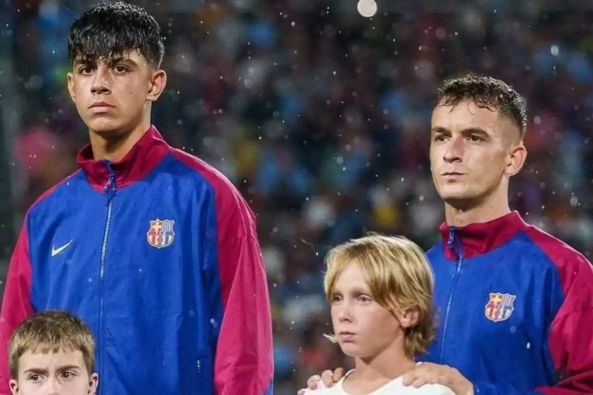 Dos jugadores de fútbol con chaquetas del FC Barcelona posan junto a dos niños en un estadio.