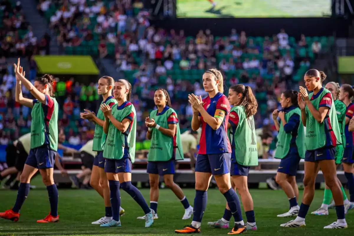 Jugadoras de fútbol con chalecos verdes aplauden y saludan al público en un estadio lleno de espectadores