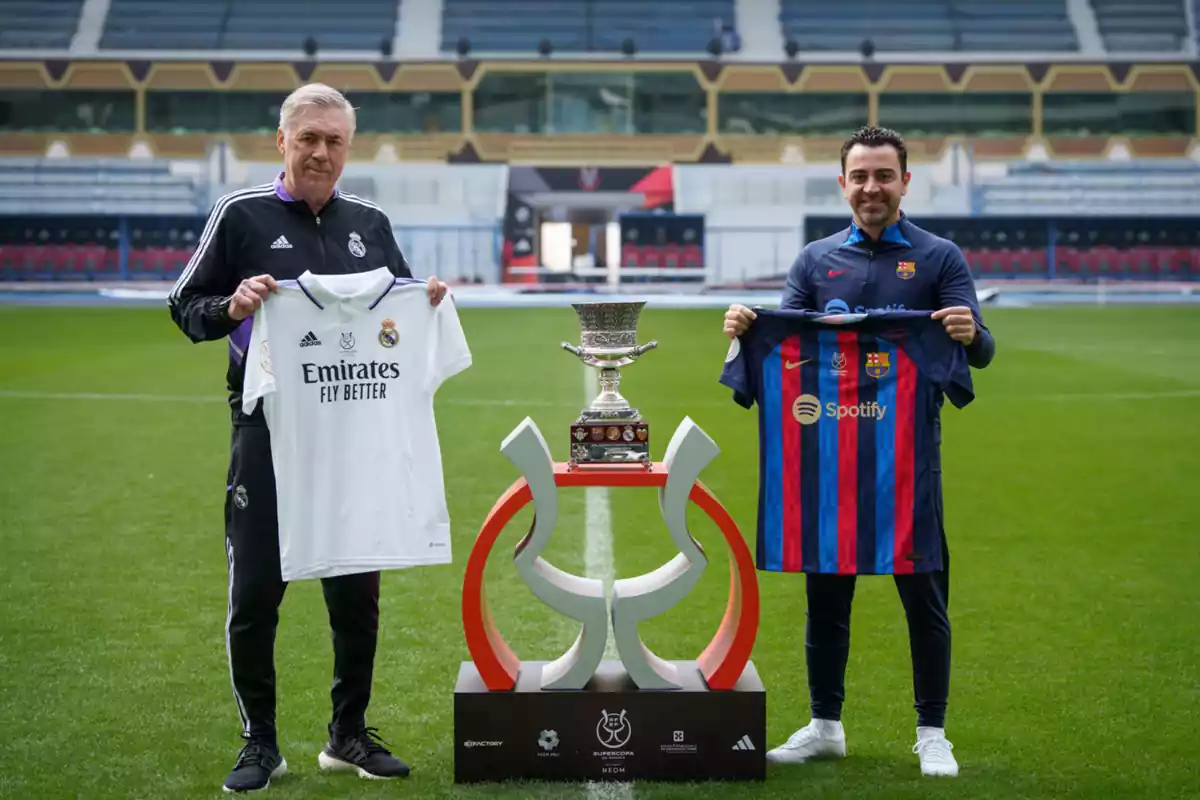 Los entrenadores de Barça y Madrid Ancelotti y Xavi, posando con la Supercopa de España y enseñando cada uno la camiseta de su equipo en un campo