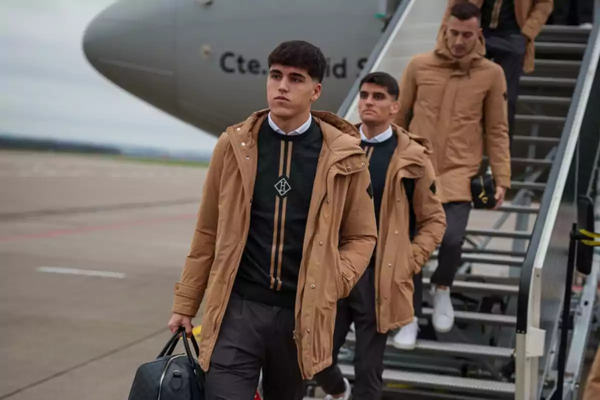 Un grupo de hombres jóvenes con abrigos marrones desciende de un avión por una escalera en un aeropuerto.