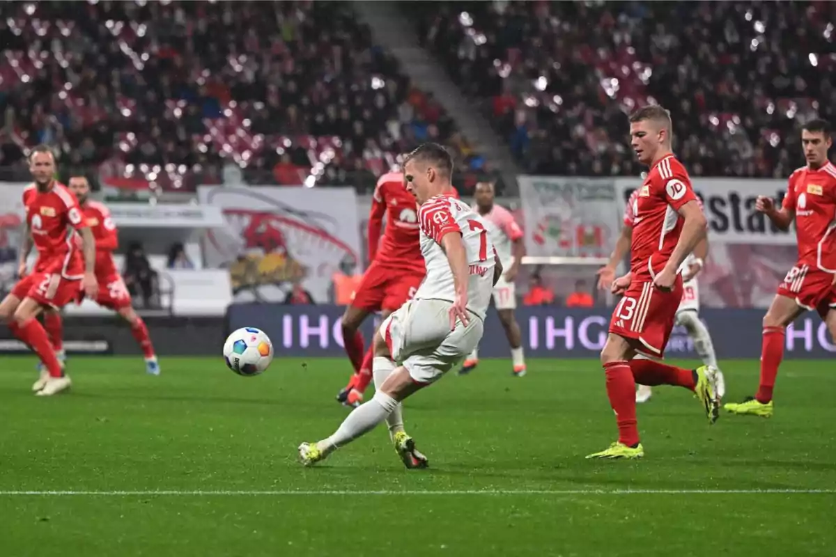 Jugadores de fútbol en acción durante un partido, con un jugador en uniforme blanco pateando el balón mientras varios jugadores en uniforme rojo lo rodean.