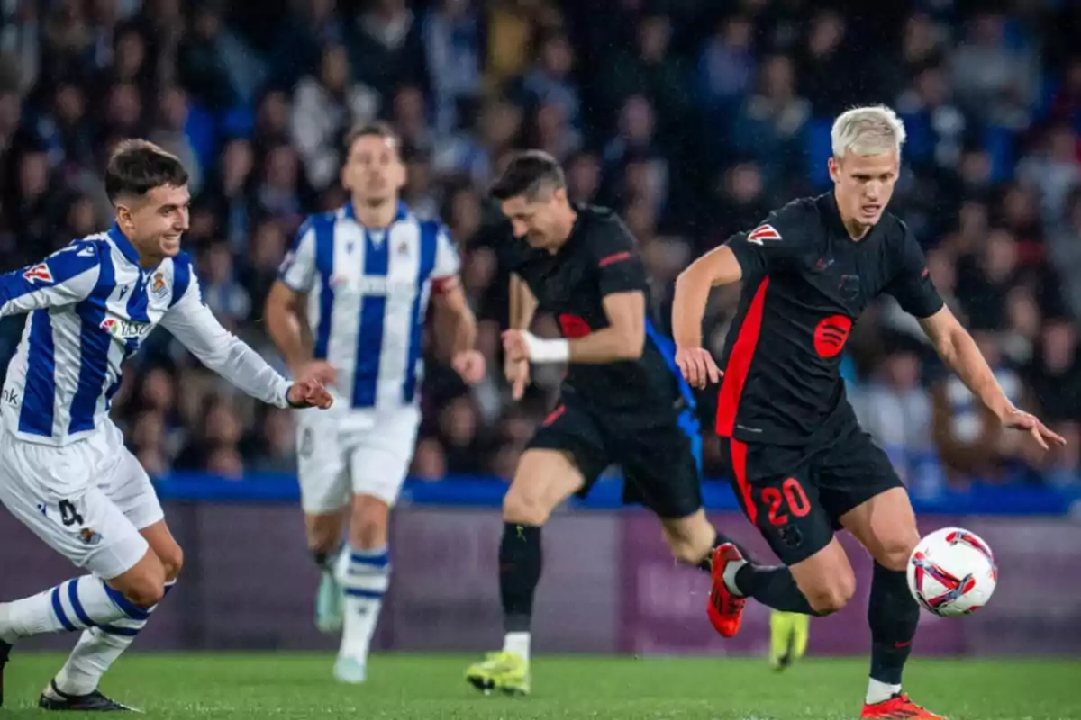 Jugadores de fútbol en acción durante un partido, uno con uniforme blanco y azul y otro con uniforme negro y rojo, en un estadio lleno de espectadores.