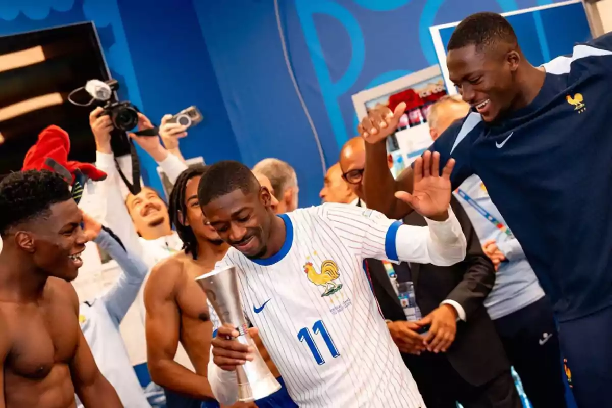 Soccer players celebrating in the locker room, one of them holding a trophy while the others applaud and smile.