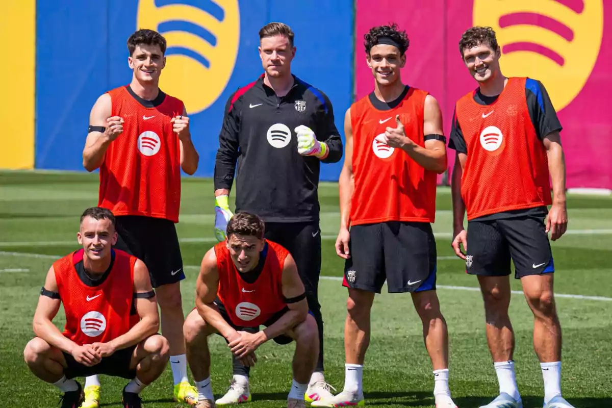Un grupo de seis jugadores de fútbol con petos rojos posan en un campo de entrenamiento con un fondo colorido. Un grupo de seis jugadores de fútbol con petos rojos posan en un campo de entrenamiento con un fondo colorido.