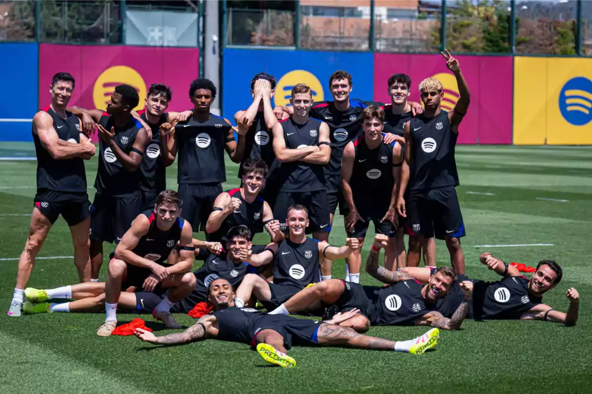 A group of players cheerfully posing on a training field, all wearing black jerseys with a visible logo, some making hand gestures while others are sitting or lying on the grass. A group of players cheerfully posing on a training field, all wearing black jerseys with a visible logo, some making hand gestures while others are sitting or lying on the grass.