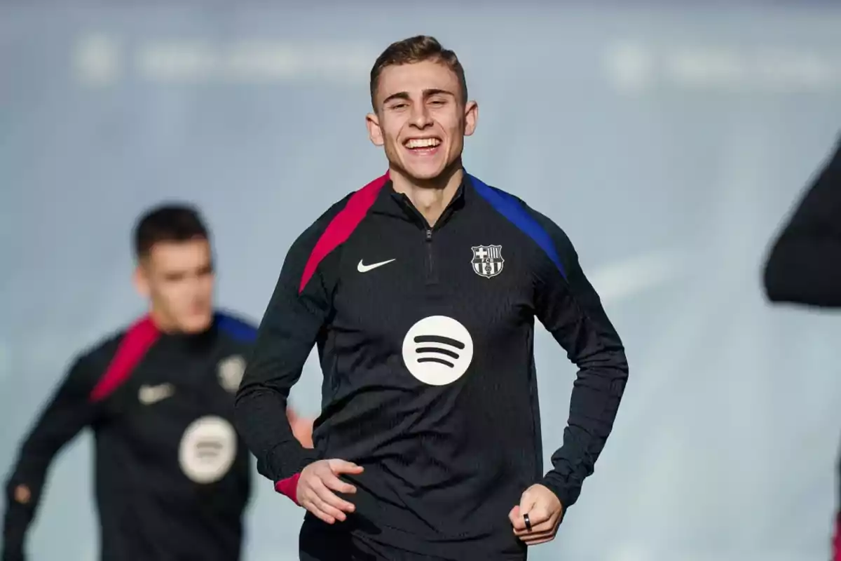 Un footballeur sourit alors qu'il court à l'entraînement, vêtu d'un uniforme noir avec des détails bleus et rouges et un logo du sponsor sur la poitrine.