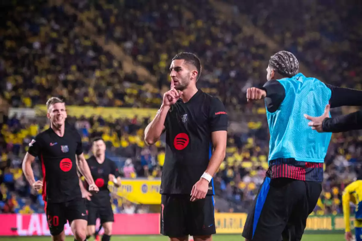 A player celebrates a goal in a stadium full of spectators, while making a shushing gesture with his finger on his lips, surrounded by teammates.