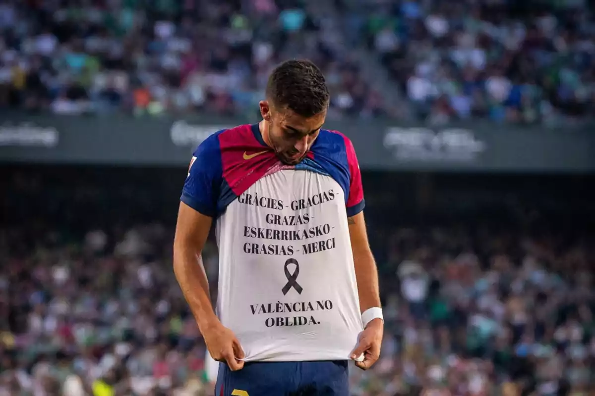 A soccer player lifts his jersey to reveal a message of thanks in several languages and a black ribbon in a stadium full of spectators.