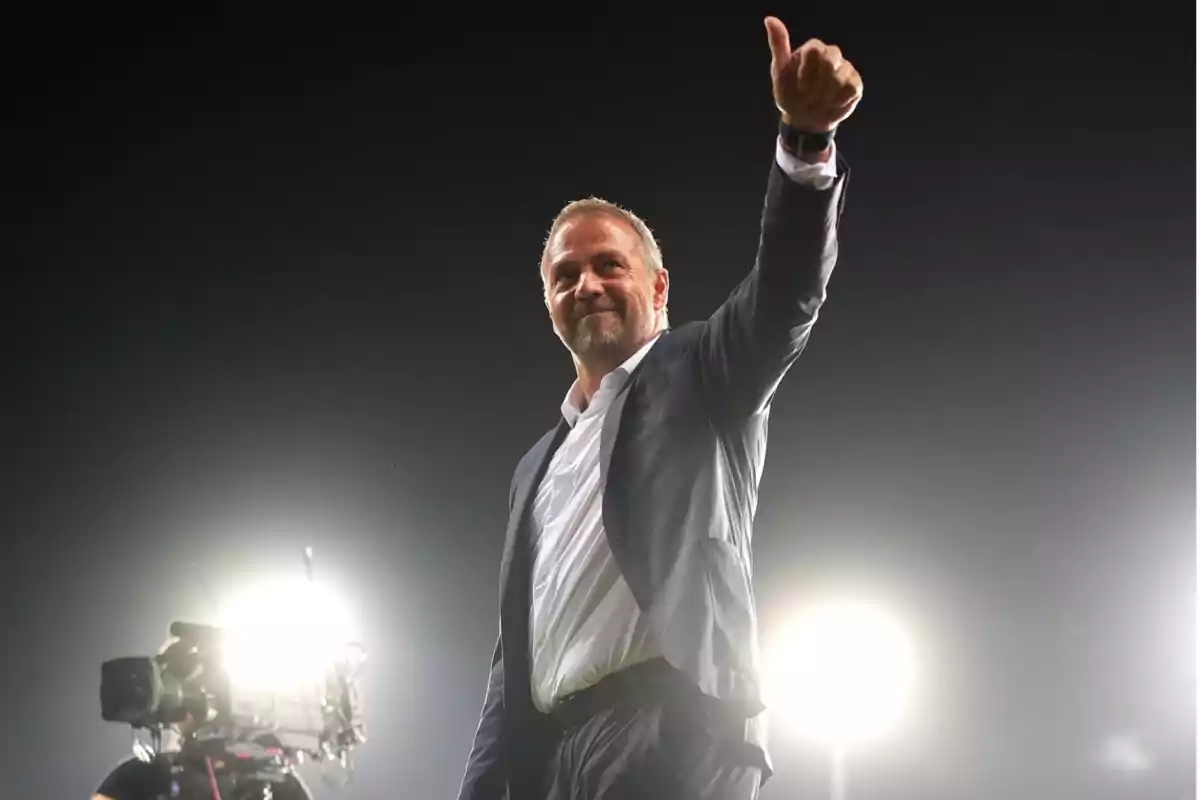 A man in a suit gives a thumbs up with bright lights in the background.