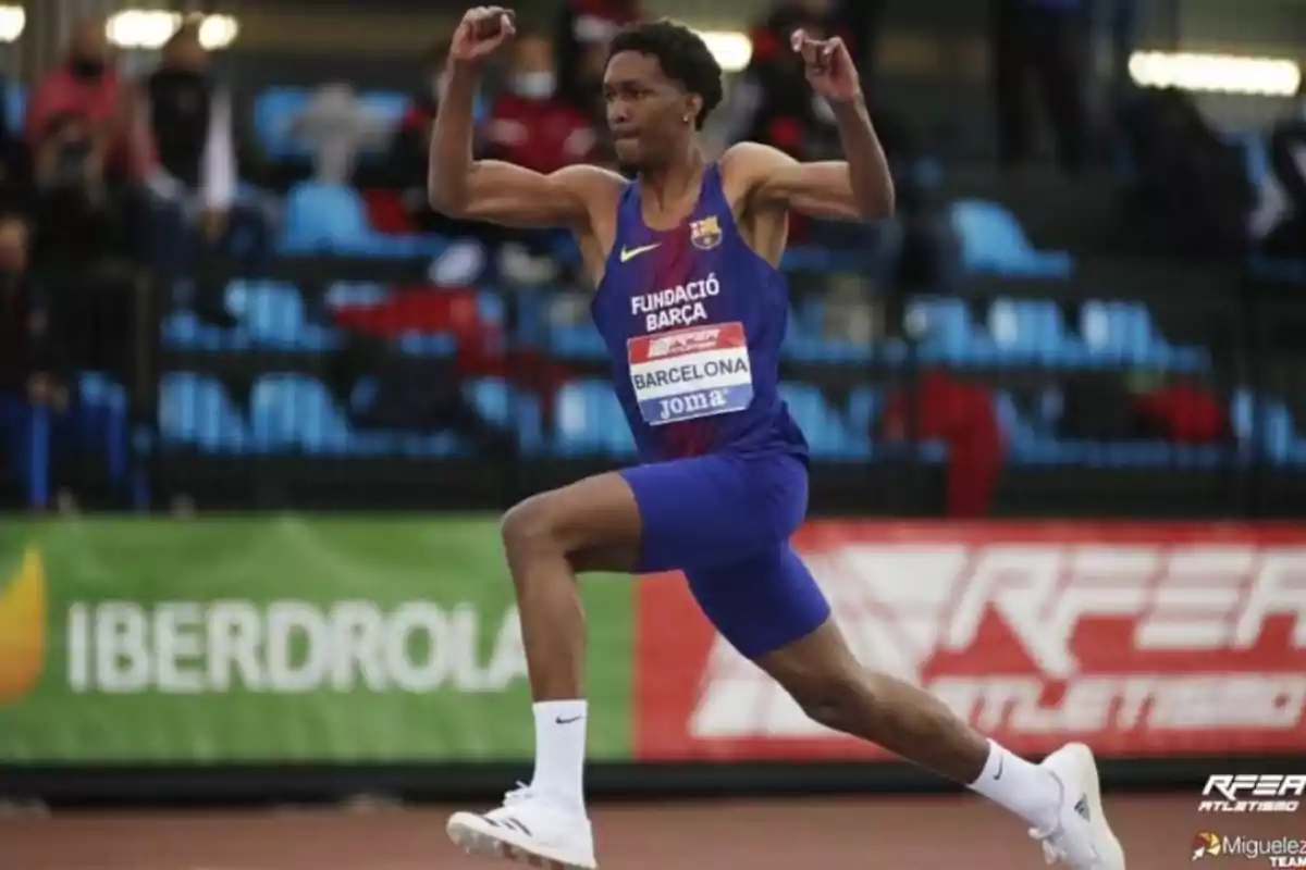 Atleta en plena competencia de salto de longitud, vistiendo un uniforme azul con el logo de la Fundació Barça y el nombre de Barcelona en el pecho, en un estadio con público de fondo.