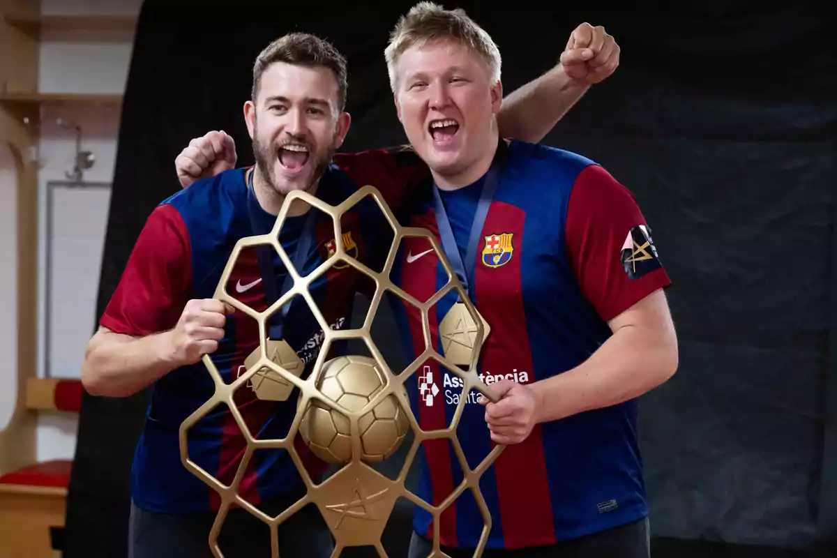 Dos hombres sonrientes con camisetas del FC Barcelona sostienen un trofeo dorado con forma de balón de fútbol y una estructura hexagonal.