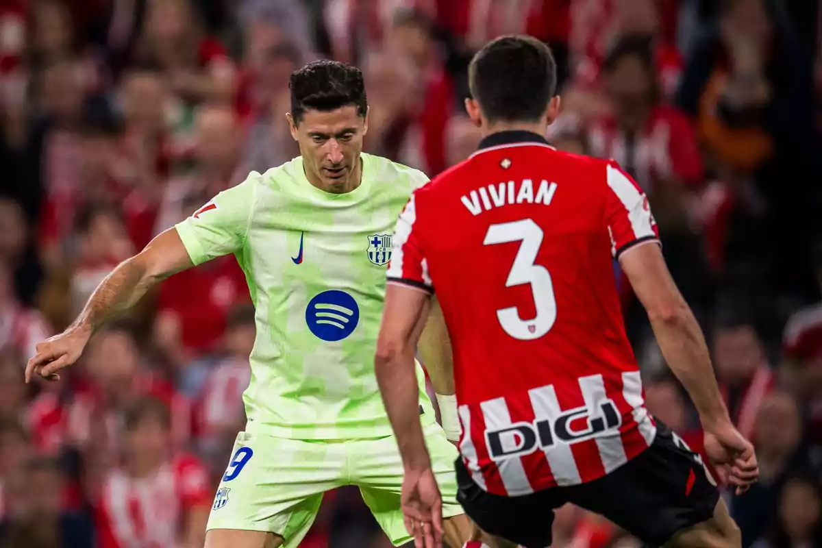 Dos jugadores de fútbol en acción durante un partido uno con uniforme verde claro y el otro con uniforme rojo y blanco a rayas Dos jugadores de fútbol en acción durante un partido uno con uniforme verde claro y el otro con uniforme rojo y blanco a rayas