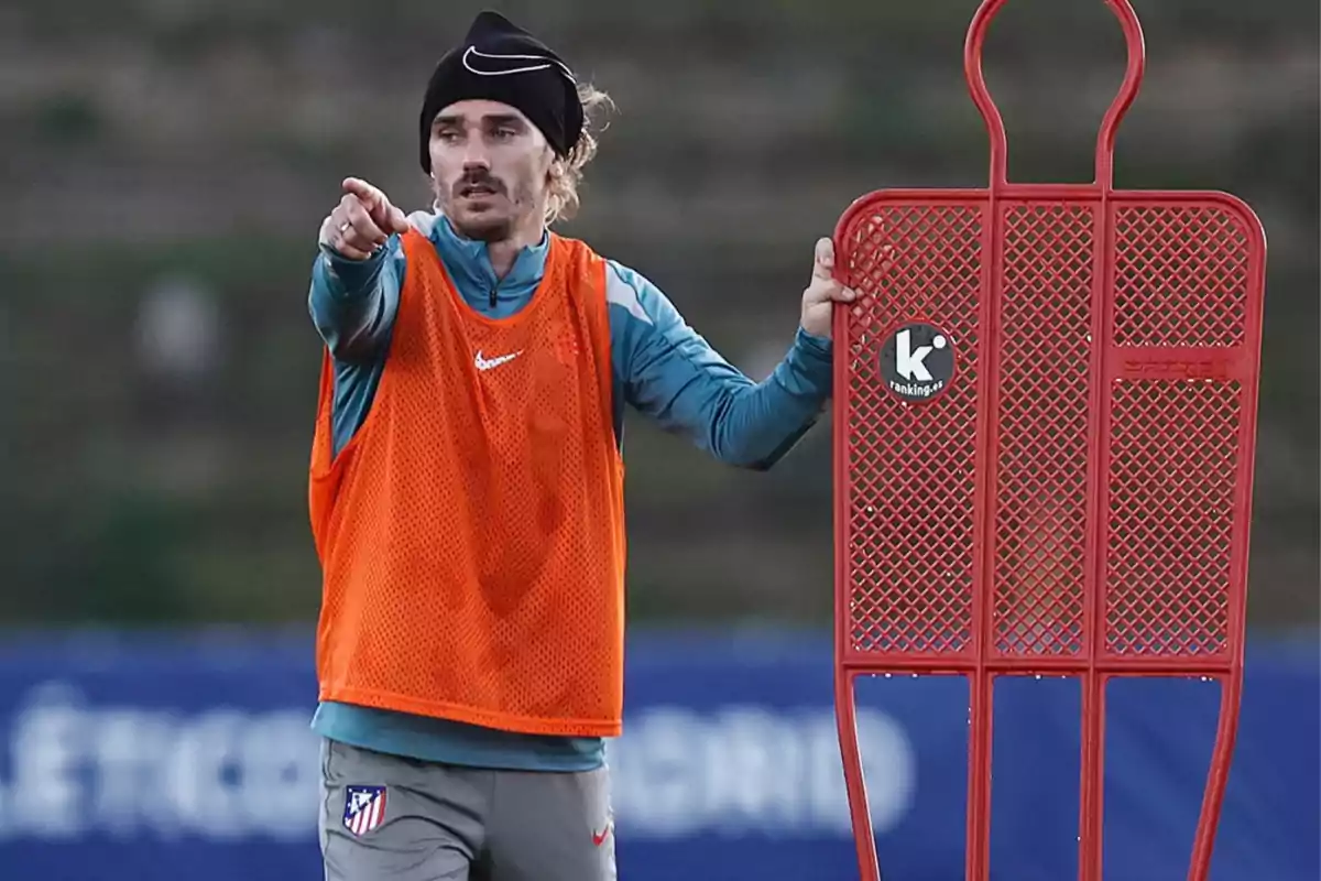 A soccer player in an orange bib and black cap points while holding a red training dummy on a field. A soccer player in an orange bib and black cap points while holding a red training dummy on a field.