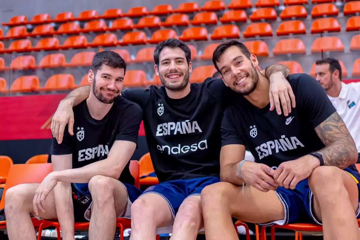 Tres jugadores de baloncesto del equipo de España sentados en las gradas de un estadio, sonriendo y abrazados.