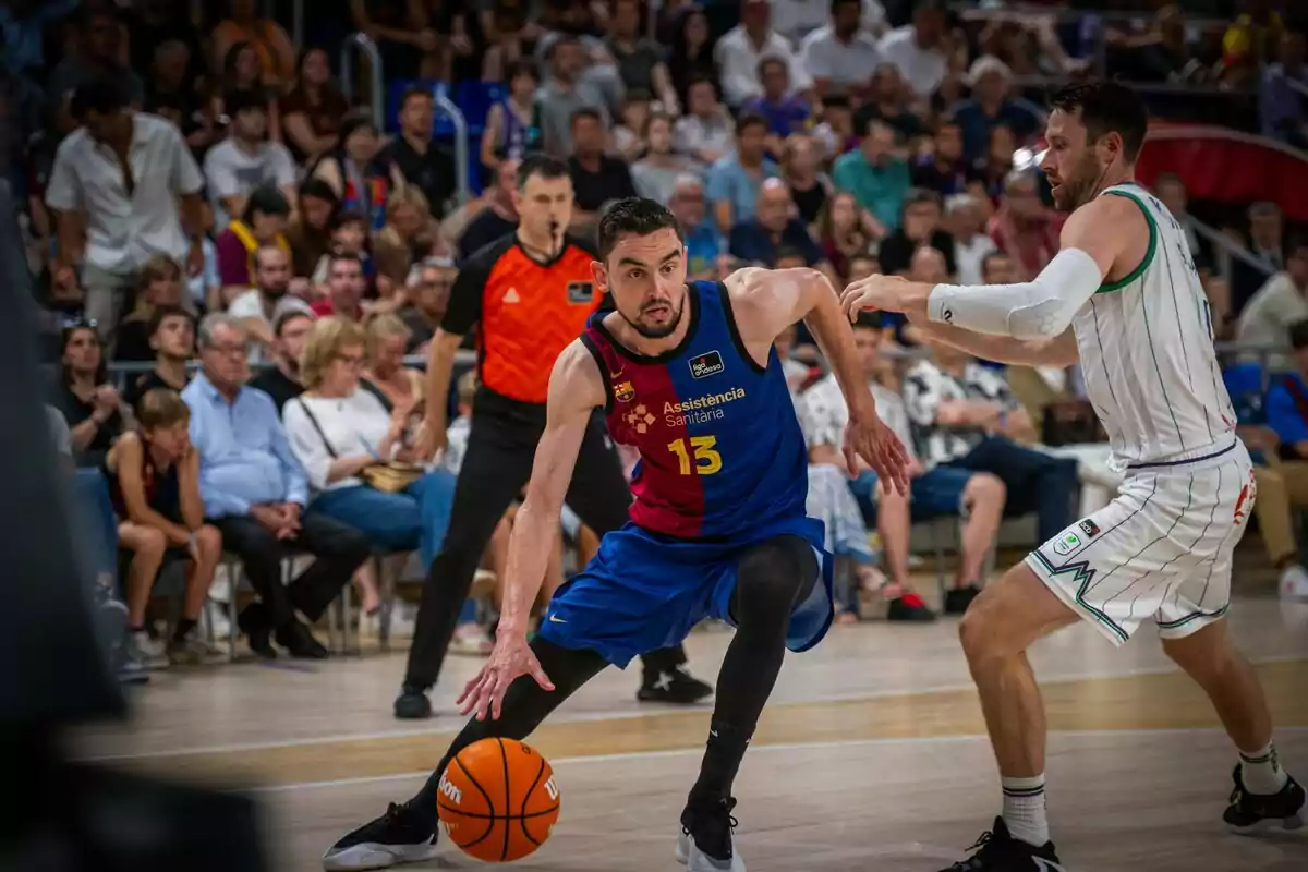 Jugador de baloncesto con uniforme azul y rojo dribla el balón mientras es defendido por un rival con uniforme blanco durante un partido con público y un árbitro al fondo