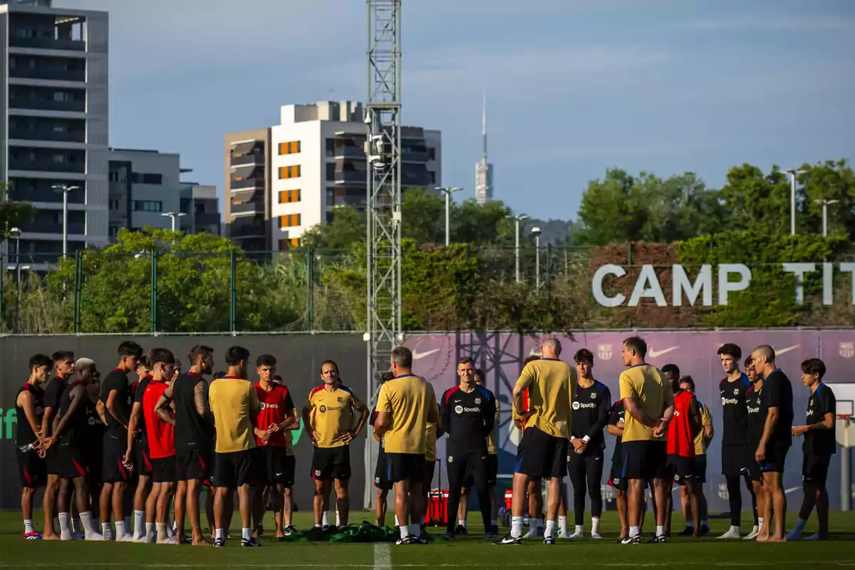 Un grupo de jugadores y entrenadores se reúne en un campo de entrenamiento al aire libre con edificios y vegetación en el fondo.