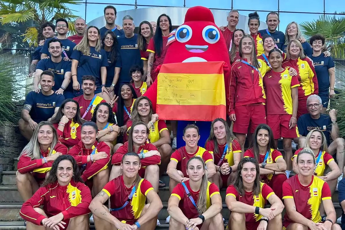 Un grupo de personas, algunas vestidas con uniformes deportivos rojos y amarillos, posan para una foto junto a una mascota roja con ojos grandes que sostiene una bandera de España firmada.