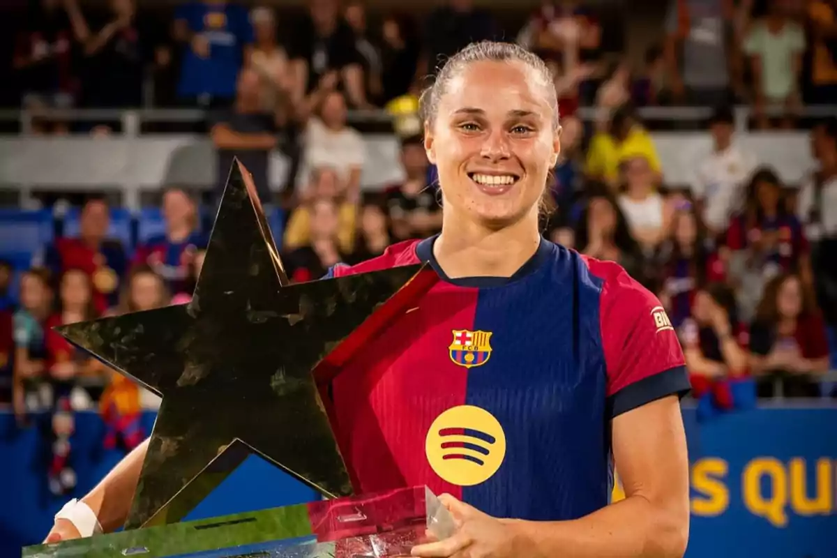 Una jugadora de fútbol sonríe mientras sostiene un trofeo en forma de estrella, vistiendo el uniforme del FC Barcelona, con una multitud de personas en el fondo.