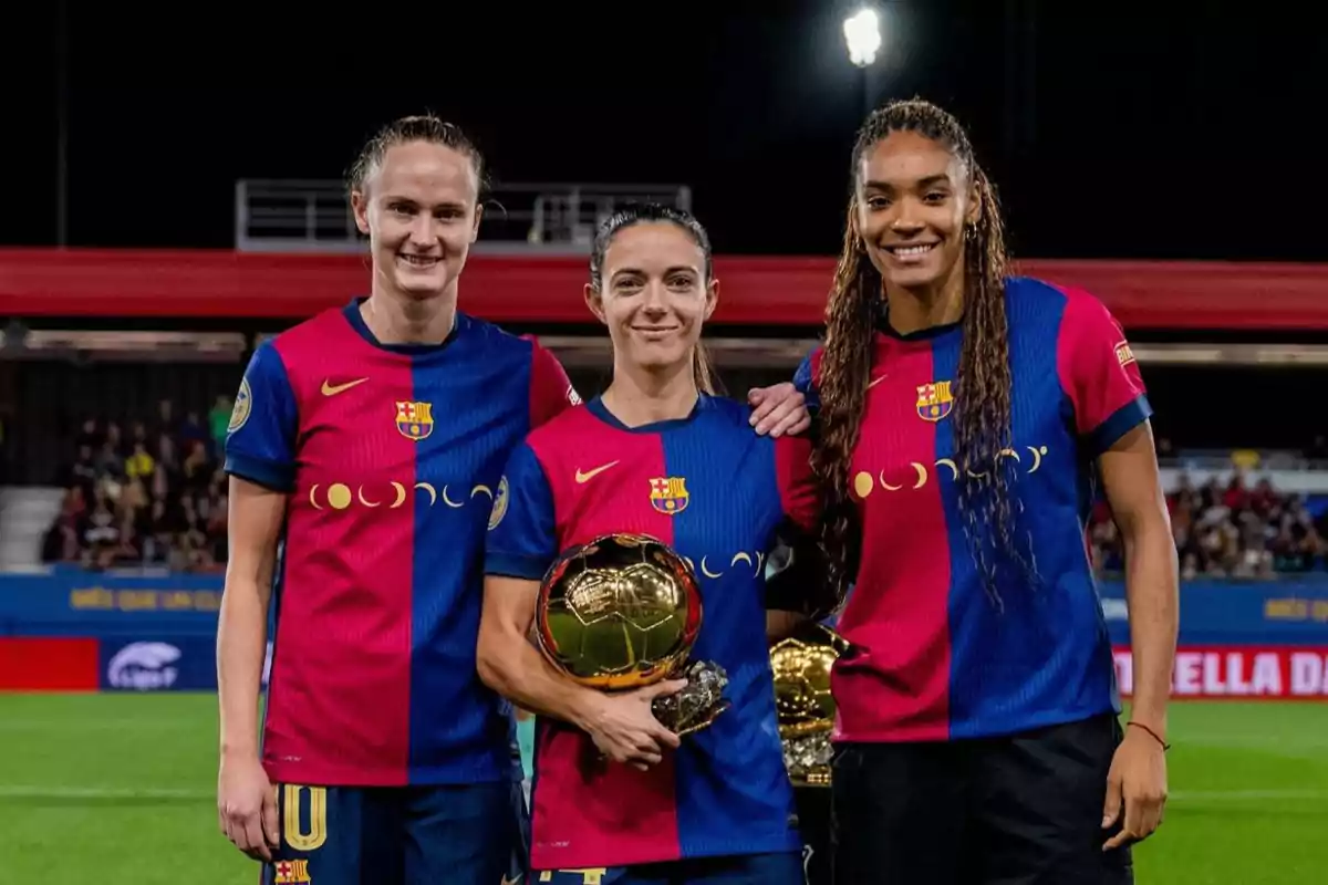 Tres jugadoras de fútbol del FC Barcelona posan en el campo, una de ellas sostiene un trofeo dorado.