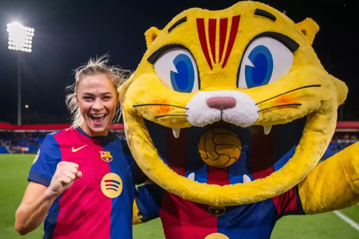 Una jugadora de fútbol sonriente junto a una mascota con uniforme del FC Barcelona en un estadio iluminado.