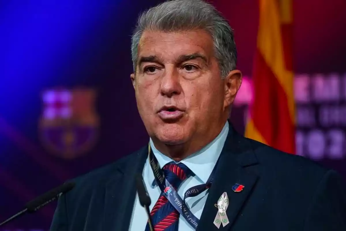 A man in a suit and tie speaks at a podium against a blue and red background.