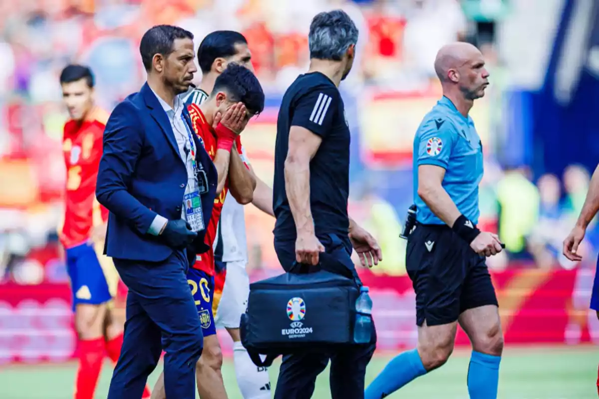 Un jugador de fútbol con la camiseta de España se cubre el rostro mientras es acompañado por personal médico y un árbitro durante un partido.