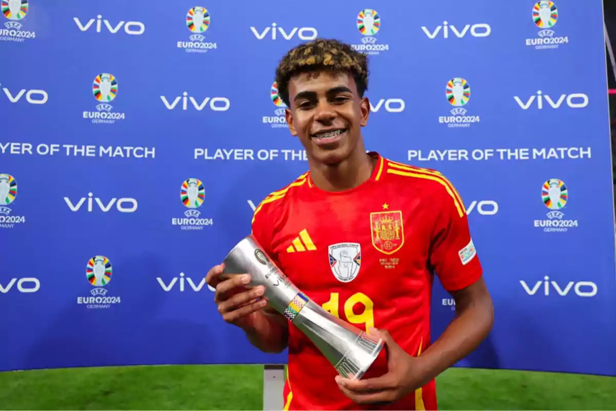 Un jugador de fútbol con la camiseta de la selección española sostiene un trofeo de "Player of the Match" frente a un fondo azul con logotipos de "vivo" y "EURO 2024".